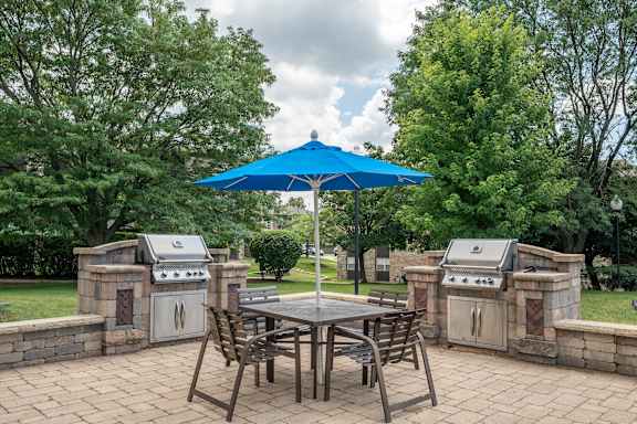 A blue umbrella is over a table with chairs and two grills in the background.