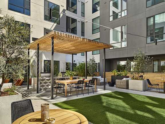 A patio with a table and chairs is surrounded by a grassy area and a building with large windows.