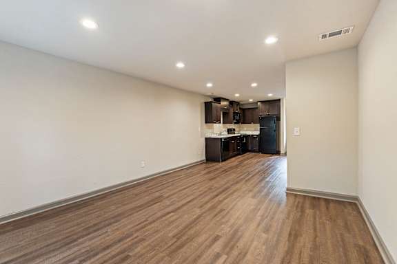 A room with wooden flooring and a kitchen area in the background.