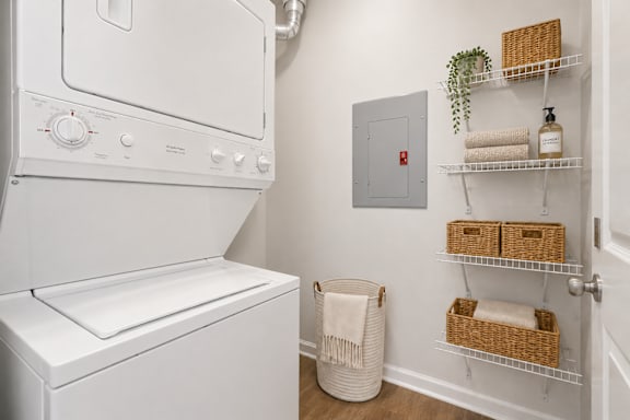 A white dryer is in a small laundry room with a basket and towels on the shelf.