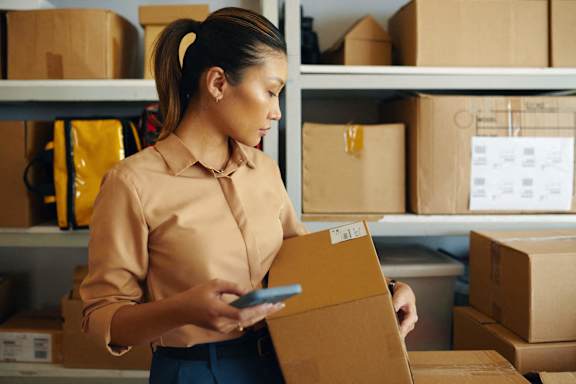 A woman in a beige shirt is holding a box and a smartphone in a storage room.
