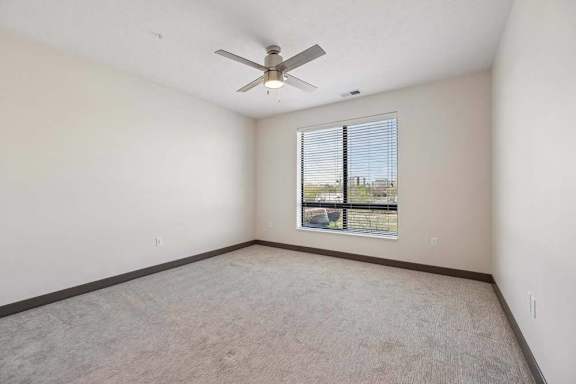 Carpeted bedroom featuring a ceiling fan and a wide window with blinds overlooking the neighborhood.