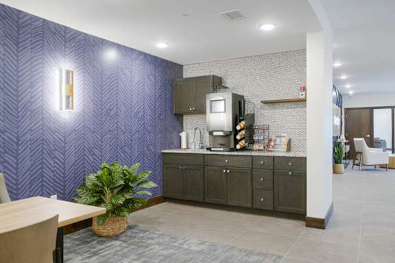 Resident coffee bar with dark cabinetry, countertop coffee machine, and open shelving, accented by patterned wallpaper, tile backsplash, and adjacent lounge seating area.