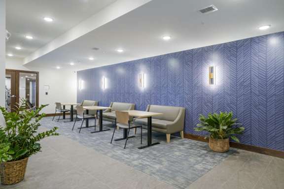 Cozy resident lounge seating area with small tables, upholstered chairs and bench seating, patterned accent wall, wall sconces, and indoor plants in a modern apartment community.