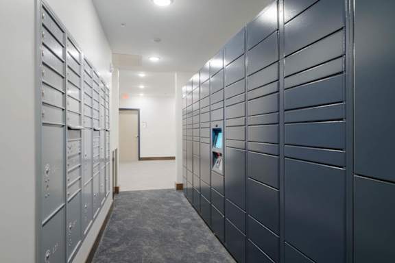 package area with wall-mounted mailboxes and electronic parcel lockers, featuring carpet flooring, bright lighting, and a clean, modern apartment community interior.