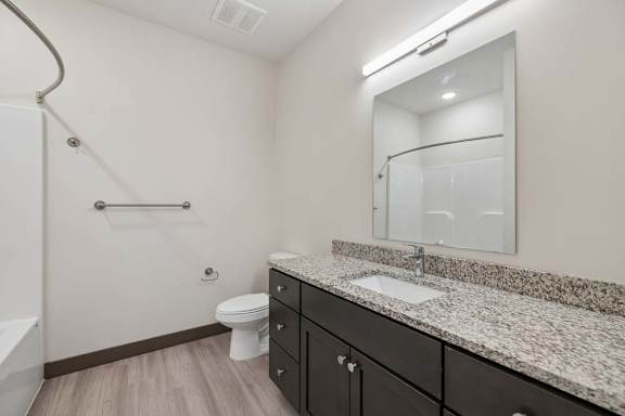 Apartment bathroom with granite countertop, dark vanity, large mirror, toilet, and tub-shower combination.