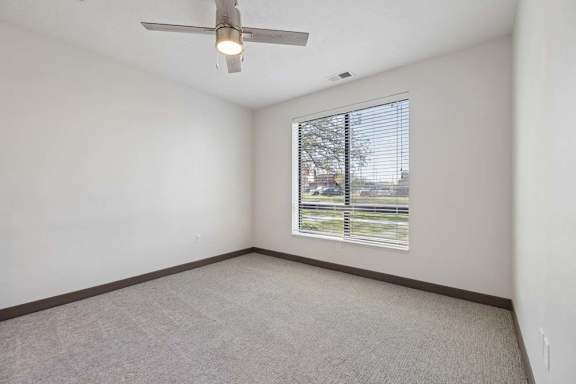 Empty apartment bedroom with neutral carpeting, a ceiling fan, and a large window with blinds overlooking the street.