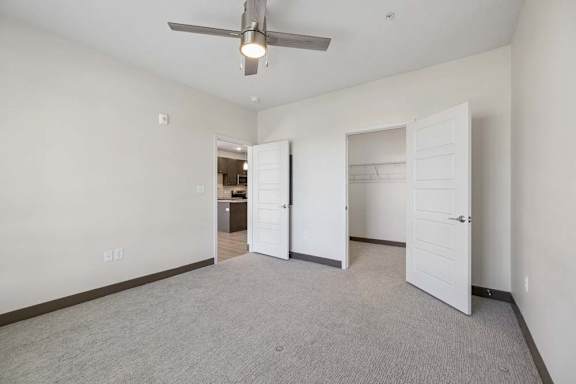 Carpeted apartment bedroom with a ceiling fan, open closet with shelving, and doorway leading to the kitchen.