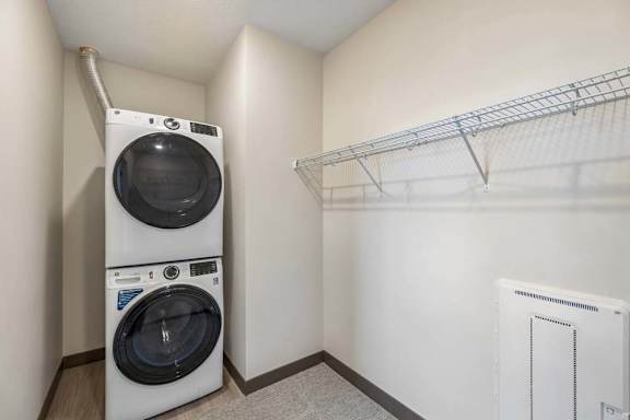 Apartment laundry and storage area with a stacked washer and dryer, carpeted floor, and wall-mounted wire shelving.