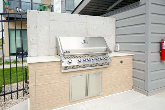 Outdoor grilling station with built-in stainless steel grill, countertop and storage, set within a landscaped apartment community patio area near building exterior.