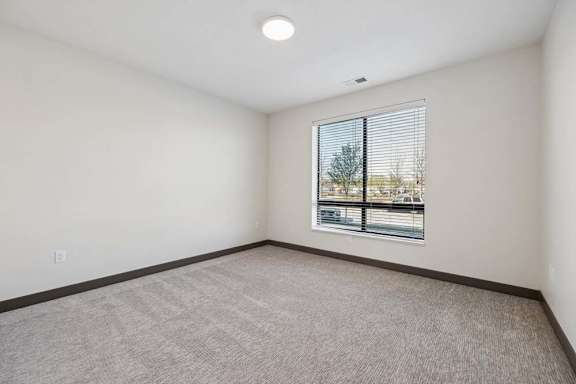 Carpeted bedroom with neutral finishes and a large window with horizontal blinds overlooking the street.