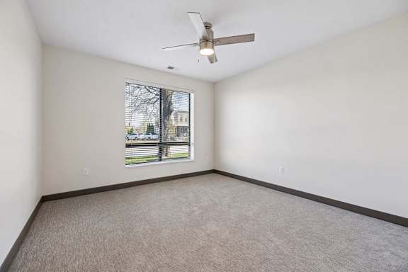 Bright, empty apartment bedroom with neutral carpet, a large window with blinds, and a modern ceiling fan with light.