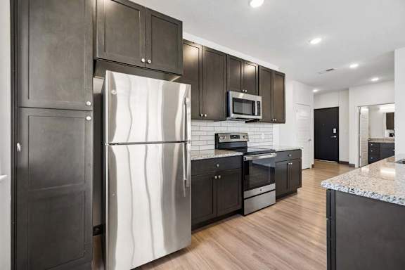 Kitchen view featuring stainless steel appliances, dark wood cabinetry, granite surfaces, and an open connection to the entry hallway.