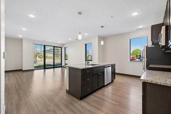 Open-concept kitchen and living area with a granite island, pendant lighting, large windows, and glass doors opening to the outdoors.