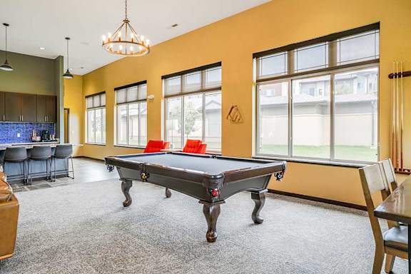 A lounge with a wooden pool table with black felt in front of several large windows.