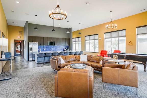 A clubhouse kitchen with large island and several bar stools, and a lounge area with a leather couch, chairs, and a pool table.