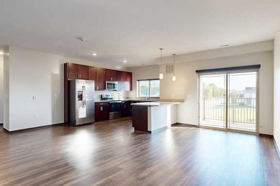 A living room with hardwood flooring, next to an open modern kitchen with dark wooden cabinets, and a sliding glass door to a balcony.