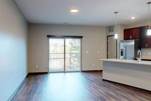 An open-concept living room with hardwood flooring and a sliding glass door to a balcony, that connects to a modern kitchen with a large central island.