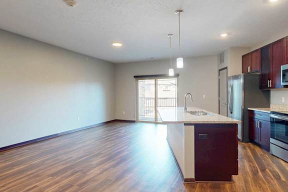 An open-concept living room with hardwood flooring and a sliding glass door to a balcony, that connects to a modern kitchen with a large central island.