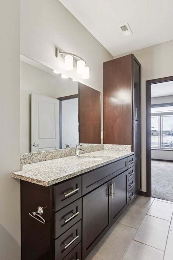A bathroom with a granite vanity, dark wooden cabinets, and a large mirror with overhead lighting.
