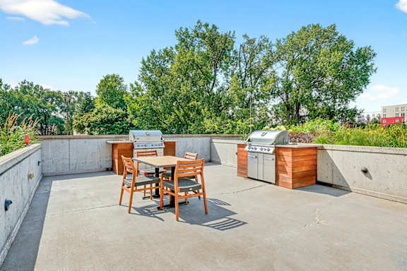 An outdoor grilling area with two built-in stainless steel grills, wooden cabinetry, and a dining table with chairs surrounded by lush greenery.