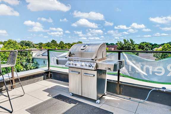 A BBQ grill is on a balcony with a view of trees and houses.