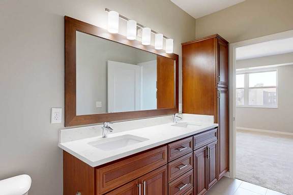 A bathroom with cherry-stained wooden cabinets, white quartz vanity, a large framed mirror and overhead lighting.