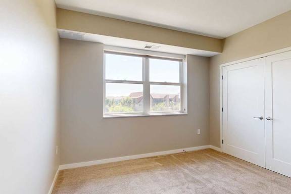 A carpeted bedroom with a alrge window and a double-door closet.