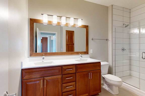 An attached bathroom with a double-sink vanity with white quartz counters, an oversized mirror, overhead lighting, and a walk-in shower with a glass door.