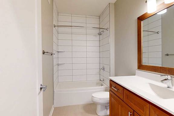 A hallway bathroom with a cherry-stained vanity sink and a tiled tub shower.