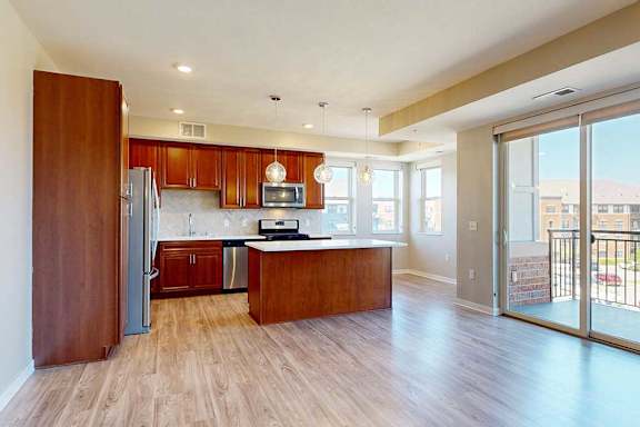 A kitchen with cherry-stained wooden cabinets, light quartz countertops, stainless steel appliances, and an island with pendant lighting in front of a spacious living room.
