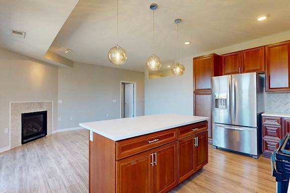 A kitchen island with cherry-stained wooden cabinets and a light quartz countertop, in front of a spacious living room with a fireplace.