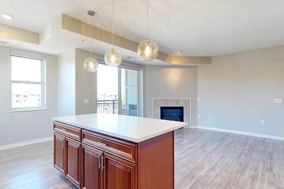 A kitchen island with cherry-stained wooden cabinets and a light quartz countertop, in front of a spacious living room with a fireplace.