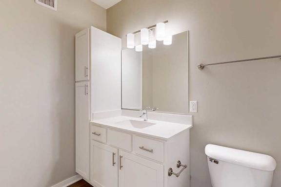 A bathroom with white cabinets, a white granite vanity, an oversized mirror with overhead lighting, and a tall storage cabinet.