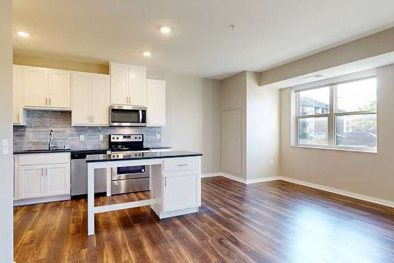 A modern kitchen with white cabinets and an island, open to a living area with large windows and hardwood-style flooring.