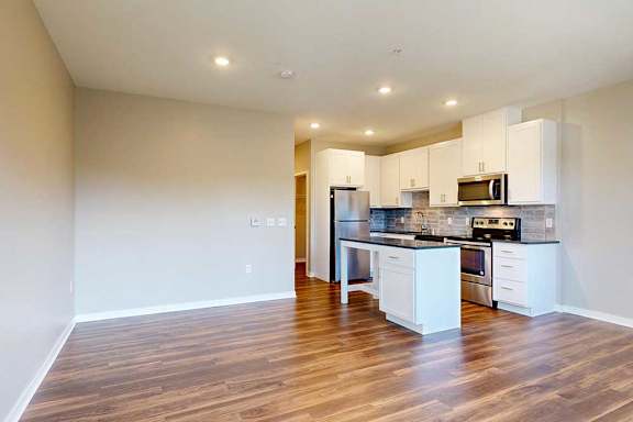 A living area with hardwood-style flooring, open to a modern kitchen with white cabinets and an island.