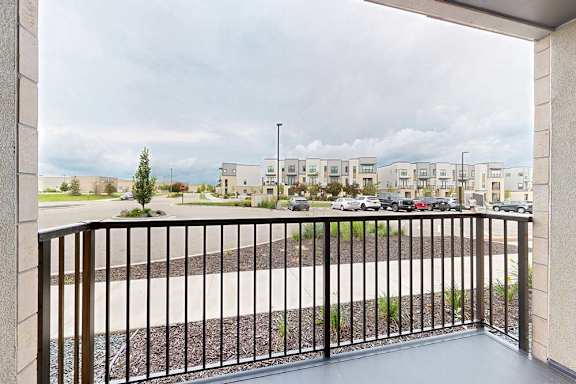 A balcony with a black railing overlooks a parking lot and apartment buildings.