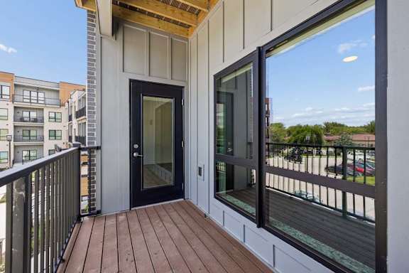 Private apartment balcony with wood-look decking, black railing, and views of the community buildings and surrounding neighborhood.