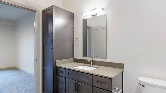 a bathroom with a wide quartz vanity sink, a slim mirror, and a tall dark wood storage cabinet