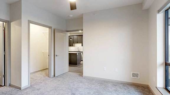 a bedroom with carpeted flooring, a door to the living room, a large window, a ceiling fan, and an L-shaped walk-in closet