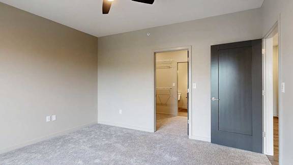 A carpeted bedroom with a walk-in closet, attached bathroom, and a door open to the living room.