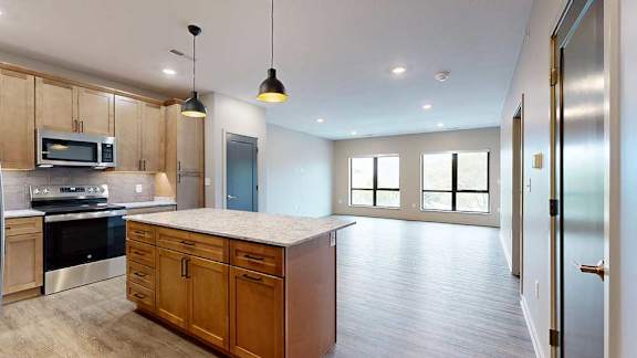 A kitchen with warm wooden cabinets and a large island, in front of a spacious living room with floor-to-ceiling windows.