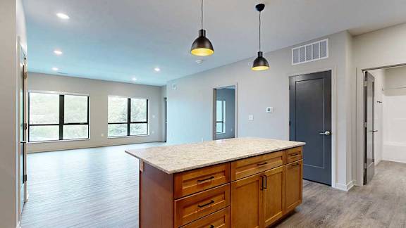 A kitchen with warm wooden cabinets and a large island, in front of a spacious living room with floor-to-ceiling windows.