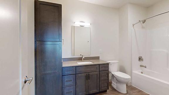 A bathroom with a quartz vanity, dark wooden cabinets, a tall storage cabinet, and a tub shower with brushed nickel fixtures.