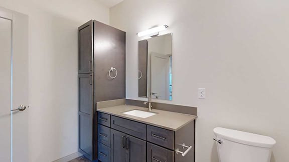 A bathroom with dark wooden cabinets, a quartz vanity sink, mirror with overhead lighting, and a tall storage cabinet.