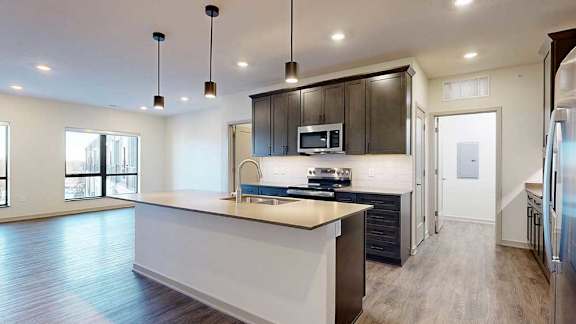 A kitchen with dark wooden cabinets and a large island, in front of a spacious living room with floor-to-ceiling windows.