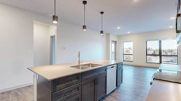 A kitchen with dark wooden cabinets and a large island, in front of a spacious living room with floor-to-ceiling windows.