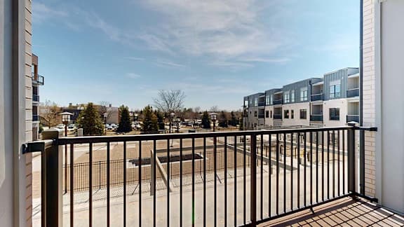 Private apartment balcony with black metal railing overlooking the community courtyard and neighboring residential buildings under a clear blue sky.