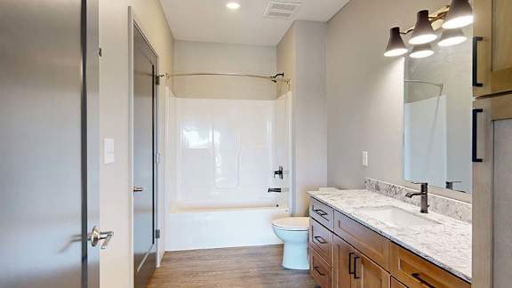 Modern apartment bathroom with a single-sink vanity, wood cabinetry, stone-look countertop, large mirror, bathtub and shower combination, and neutral finishes.