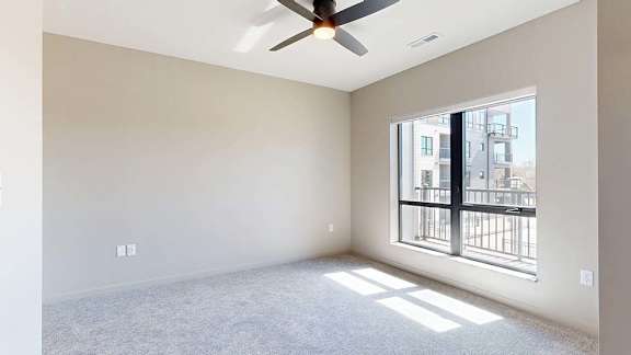 Bright apartment bedroom with soft carpeting, a large window providing natural light, a ceiling fan, neutral finishes, and views of the neighboring community buildings.